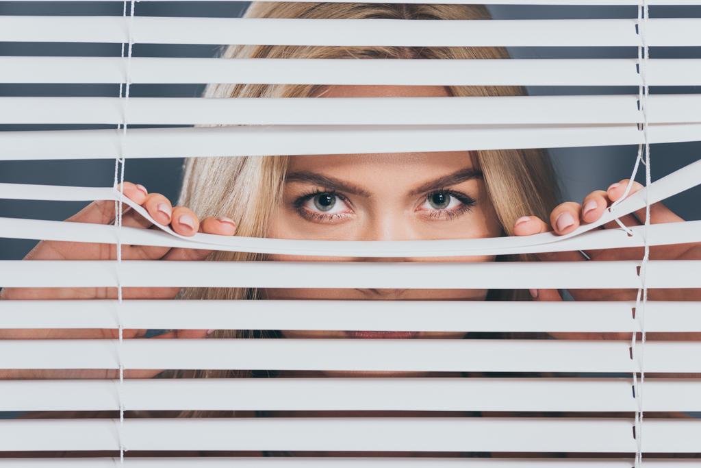 young woman looking at camera and peeking through blinds  - Photo, Image