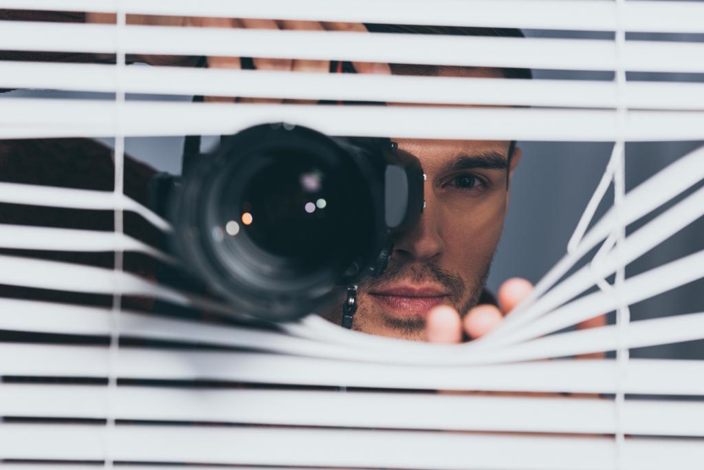 jonge man houden van de camera en de camera via blinds, wantrouwen concept kijken - Foto, afbeelding