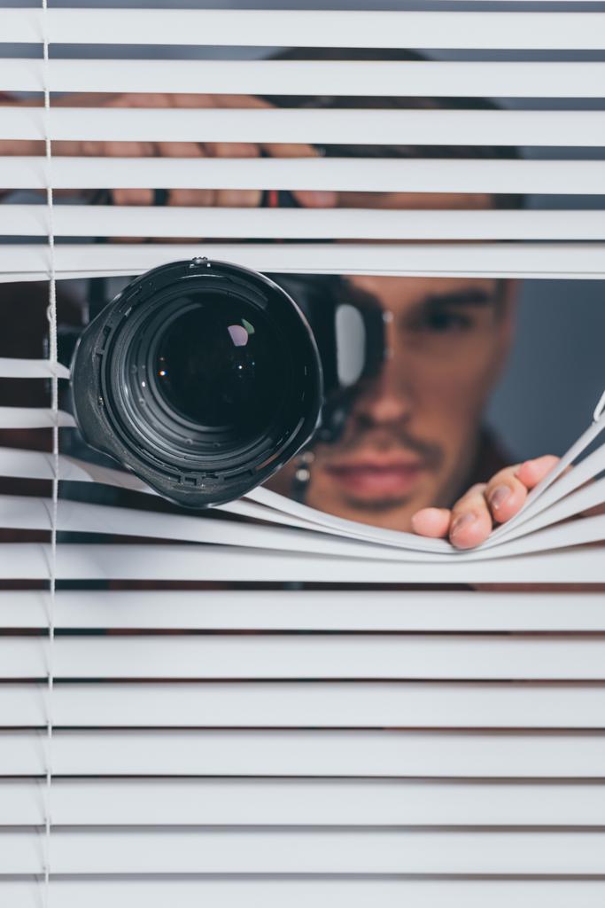 vista de cerca del joven sosteniendo la cámara y mirando a la cámara a través de persianas
 - Foto, imagen