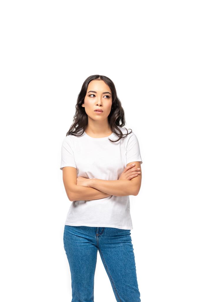 nachdenkliche Asiatin in weißem T-Shirt und blauer Jeans, die mit verschränkten Armen auf weißem Hintergrund steht  - Foto, Bild
