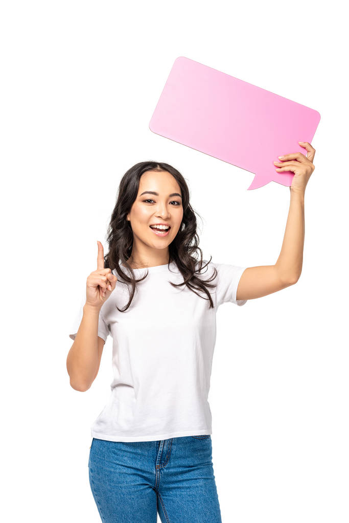 Mujer asiática sonriente en camiseta blanca y jeans azules sosteniendo la burbuja del habla y mostrando el dedo índice aislado en blanco
 - Foto, imagen