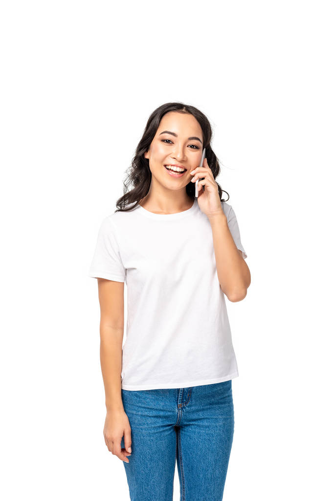 Glimlachend Aziatische meisje in wit t-shirt en spijkerbroek praten over smartphone geïsoleerd op wit - Foto, afbeelding