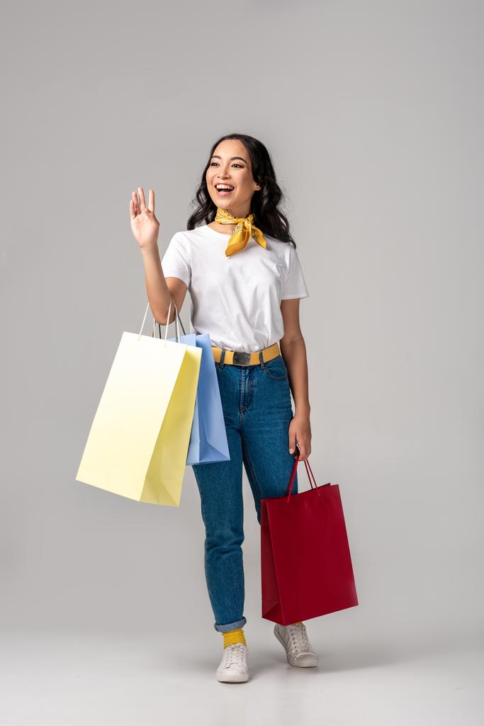 Moda vestida joven mujer asiática sosteniendo coloridas bolsas de compras y saludando con la mano levantada en gris
 - Foto, imagen