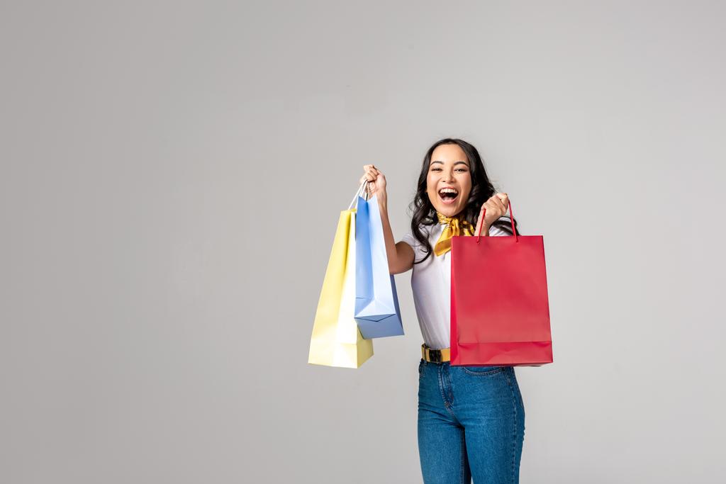Улыбающаяся азиатка, держащая красочные сумки для покупок на поднятых руках и смотрящая на камеру, изолированную на сером
 - Фото, изображение