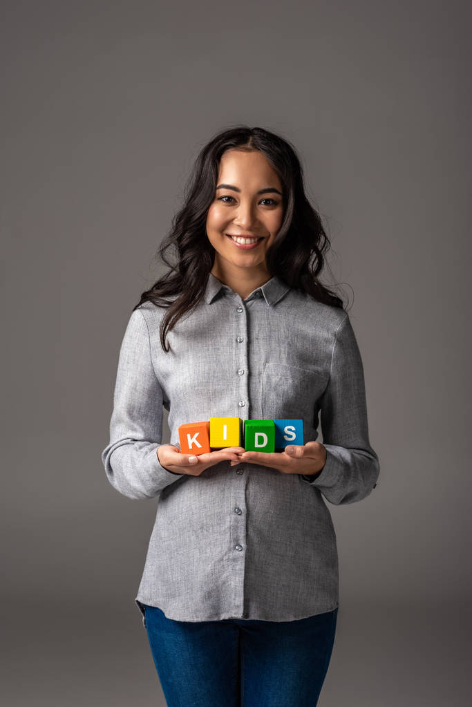 весела молода азіатська жінка тримає кубики абетки зі словом діти на сірому
 - Фото, зображення