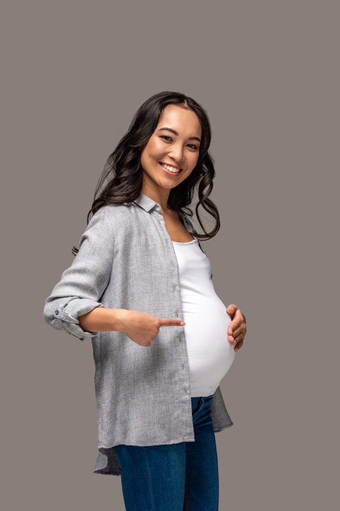 Młoda w ciąży asian Kobieta wskazując na brzuch i uśmiecha się na białym tle na szary - Zdjęcie, obraz