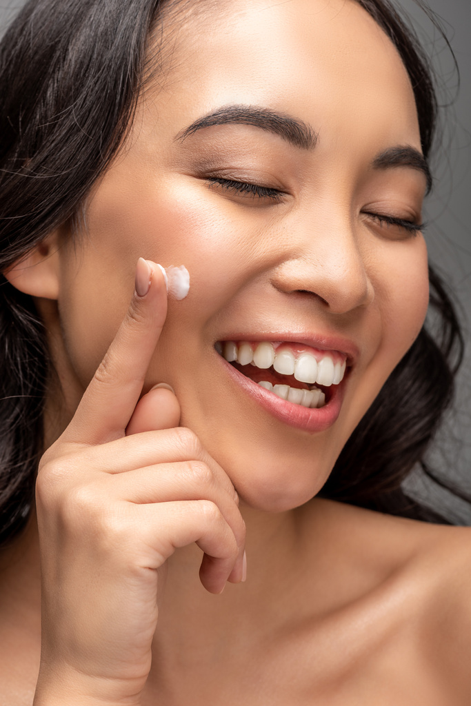 Retrato de sorrir asiático bela jovem com olhos fechados aplicando creme cosmético no rosto isolado no cinza
 - Foto, Imagem