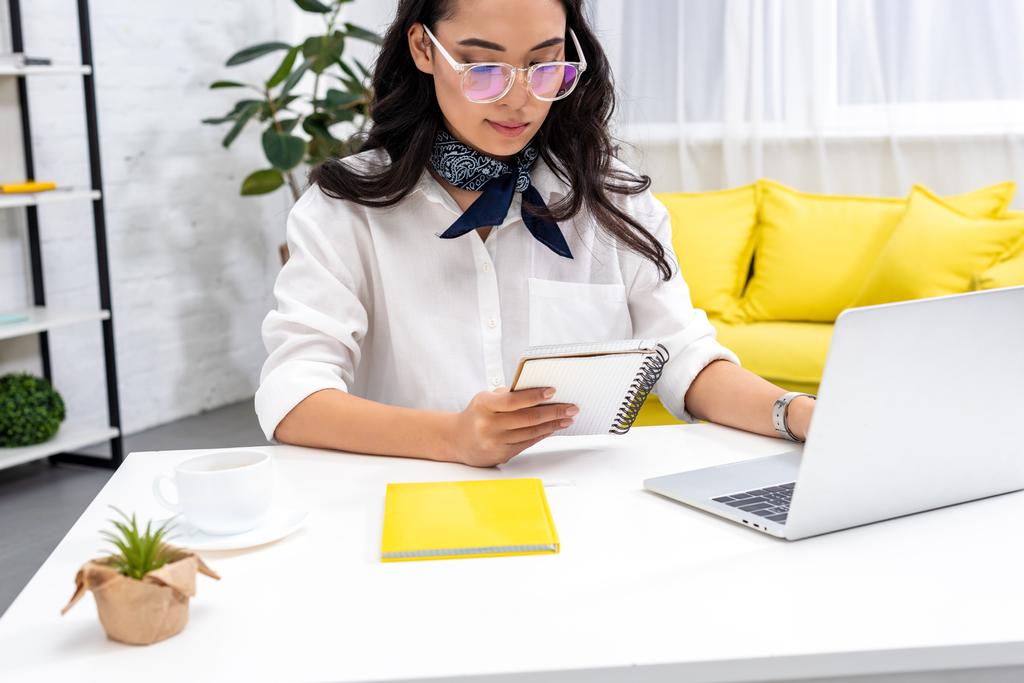 Ασιατικές αρκετά ελεύθερος επαγγελματίας σε γυαλιά χρησιμοποιούν φορητό υπολογιστή και την ανάγνωση σημειώσεις στο σημειωματάριο - Φωτογραφία, εικόνα