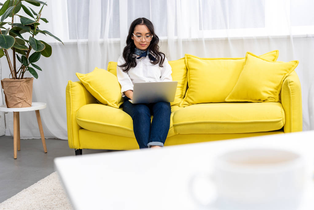 Foco seletivo de freelancer asiático sentado no sofá amarelo e usando laptop em casa
 - Foto, Imagem