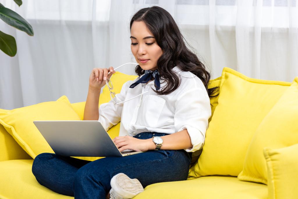 Привабливий азіатський фрілансер, використовуючи ноутбук і тримаючи окуляри в руці, сидячи зручно на жовтому дивані вдома
 - Фото, зображення