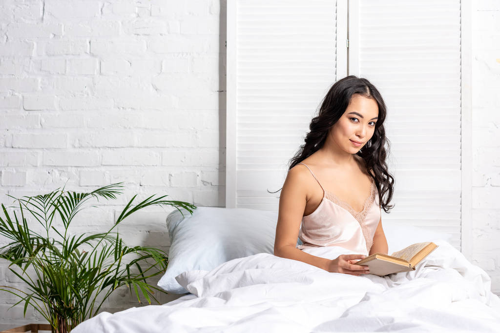 hübsche asiatische Frau im eleganten Nachthemd sitzt im Bett und hält Buch, während sie in die Kamera schaut - Foto, Bild