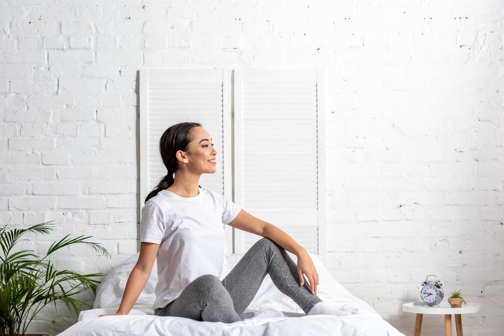 азиатская женщина в белой футболке и серых леггинсах сидит в постели и отдыхает после утренней тренировки
 - Фото, изображение