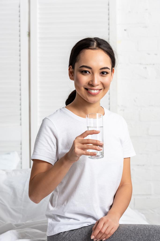 lächelnde Asiatin in weißem T-Shirt, die mit einem Glas Wasser auf dem Bett sitzt - Foto, Bild