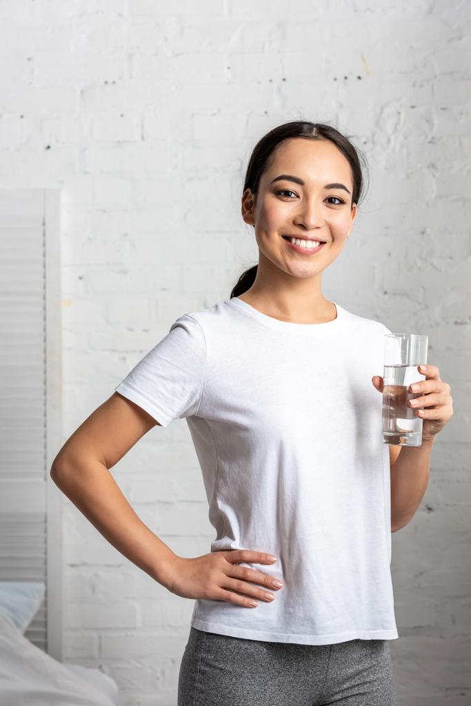 schöne junge asiatische Frau mit einem Glas Wasser steht im Schlafzimmer mit der Hand auf der Hüfte und blickt in die Kamera - Foto, Bild