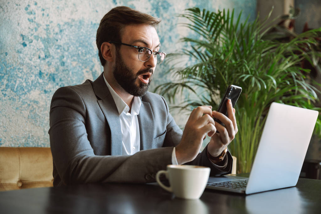 Giovane uomo d'affari scioccato vestito in giacca e cravatta che lavora su un computer portatile, seduto al caffè in casa, con il cellulare in mano
 - Foto, immagini