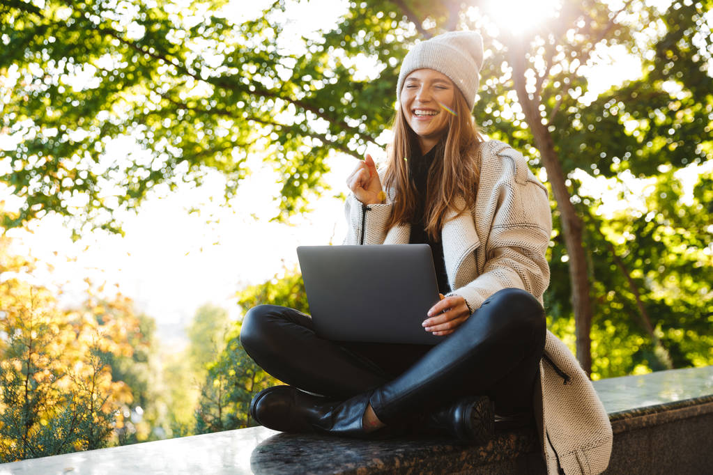 秋のコートと屋外に座って、ラップトップ コンピューターを使用して、成功を祝っての帽子に身を包んだ美しい若い女性 - 写真・画像