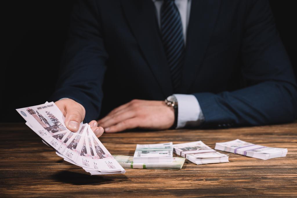 Средняя секция бизнесмена в формальной одежде держит банкноты российских рублей над деревянным столом
  - Фото, изображение