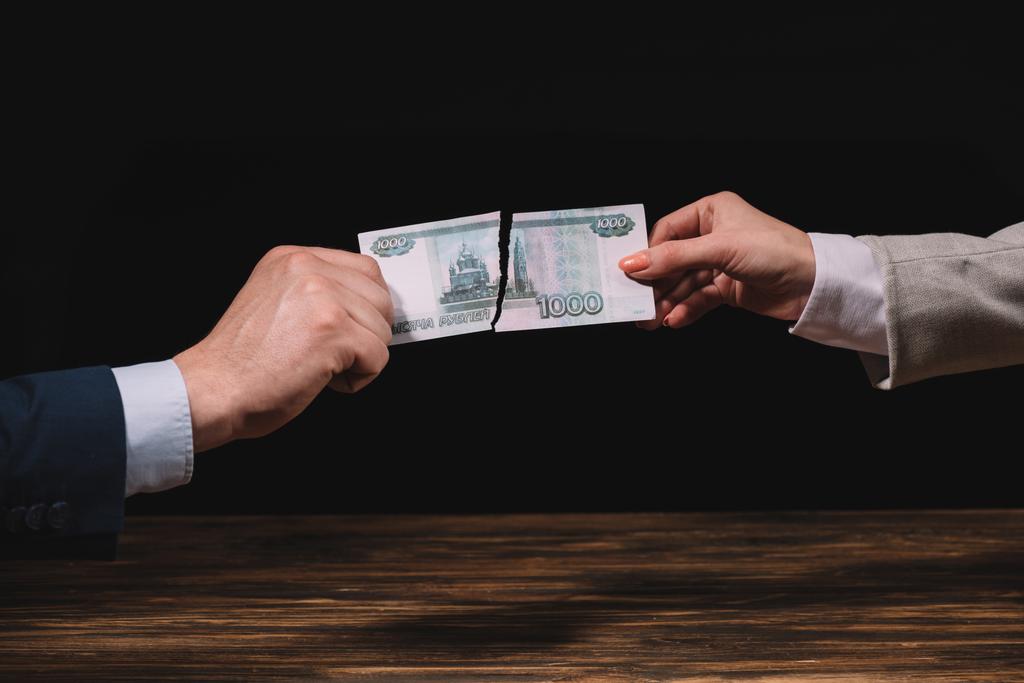 обрезанный снимок бизнесмена и предпринимательницы, разорвавшей банкноту российских рублей над деревянным столом на черном
 - Фото, изображение