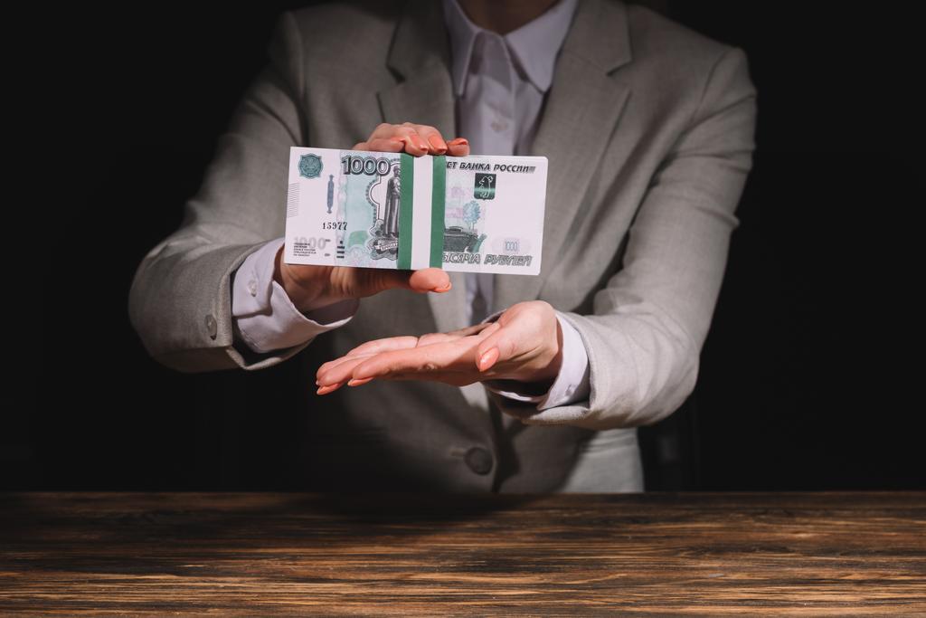 mid section of businesswoman sitting at wooden table and holding russian rubles banknotes on black  - Photo, Image