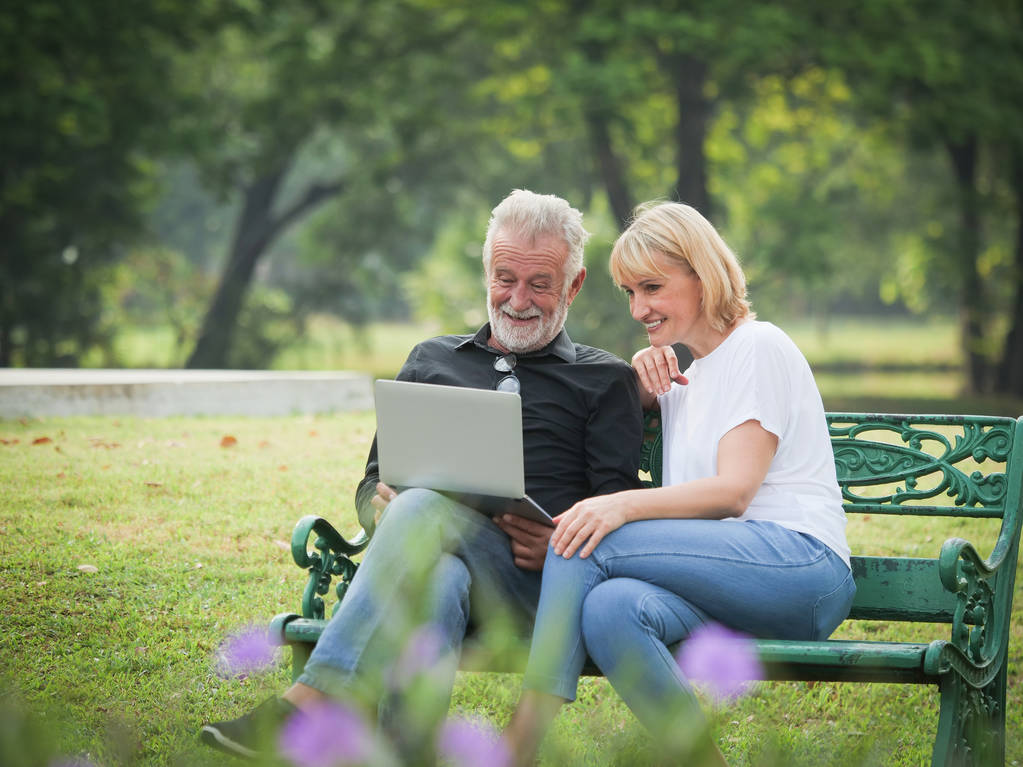 Dwa szczęśliwy seniorów emerytalne mężczyzna i kobieta siedzą i przy użyciu komputera przenośnego w parku - Zdjęcie, obraz