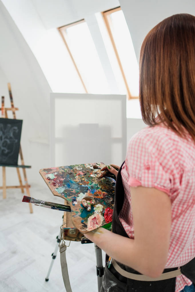 junge schöne Malerin in einem weißen Atelier zeichnet auf einer Staffelei auf Leinwand. - Foto, Bild