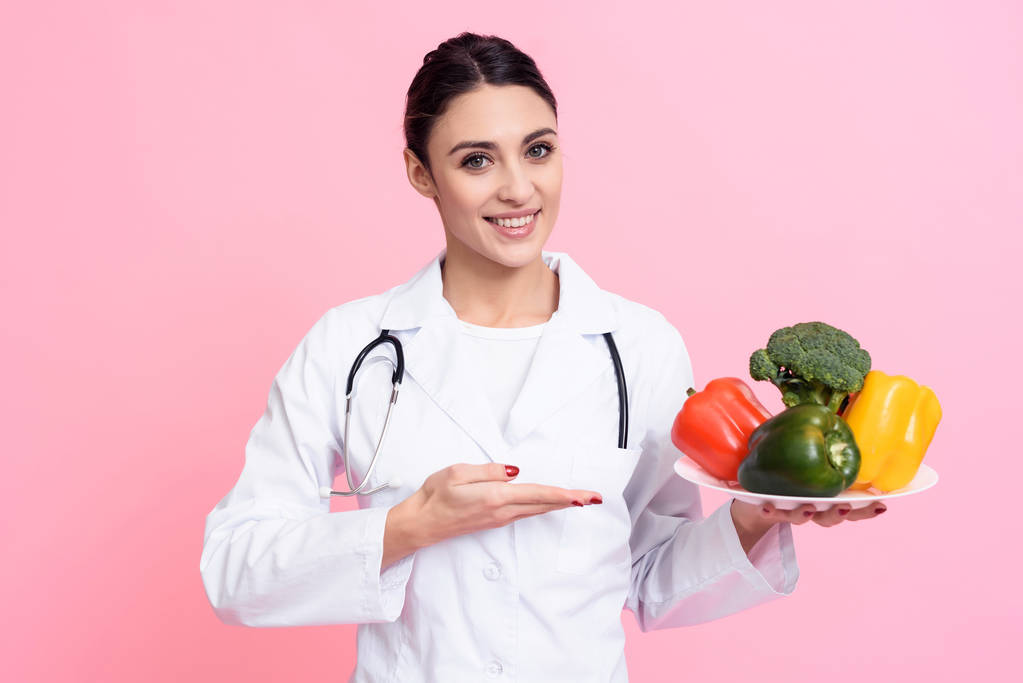 Portret van lachende vrouwelijke arts met de stethoscoop plaat van groenten geïsoleerd te houden. - Foto, afbeelding