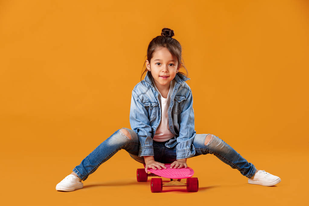 Denim içinde kaykay turuncu arka plan üzerinde şık küçük çocuk kızla - Fotoğraf, Görsel