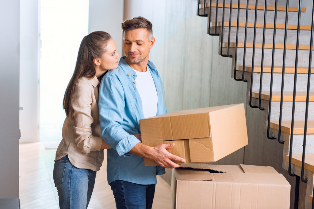 attractive woman hugging handsome husband with box in hands in new home  - Photo, Image