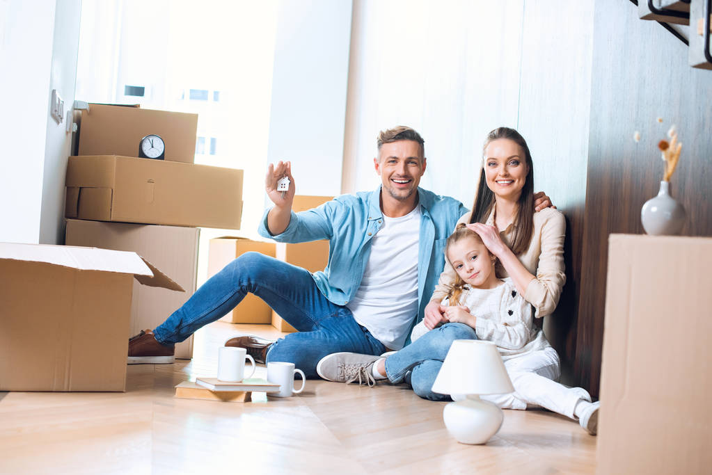 szczęśliwy człowiek siedzi na podłodze z domu w kształcie breloczek w pobliżu atrakcyjnych żona i córka  - Zdjęcie, obraz