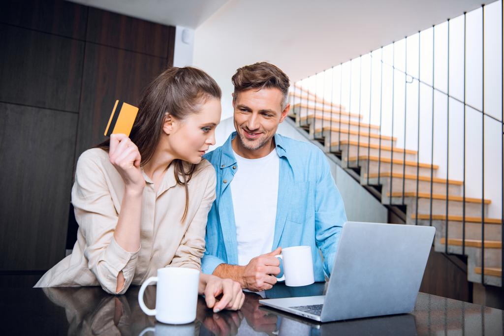 щасливий чоловік дивиться на привабливу дружину, що тримає кредитну картку біля ноутбука
  - Фото, зображення