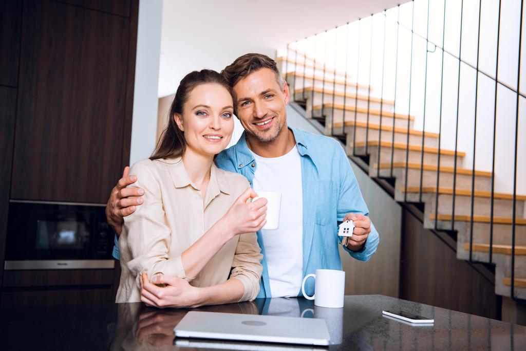 attraktive Frau, die eine Tasse Kaffee hält und mit ihrem fröhlichen Mann steht, der einen Schlüsselanhänger in Form eines Hauses hält  - Foto, Bild