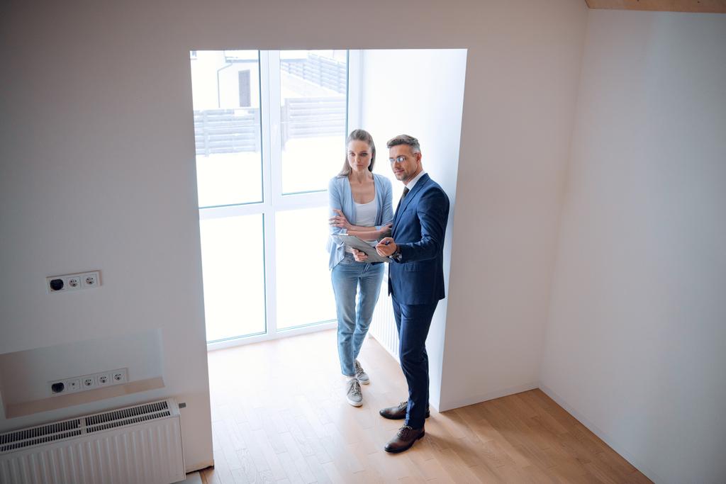 handsome broker holding clipboard and standing with beautiful woman in room  - Photo, Image