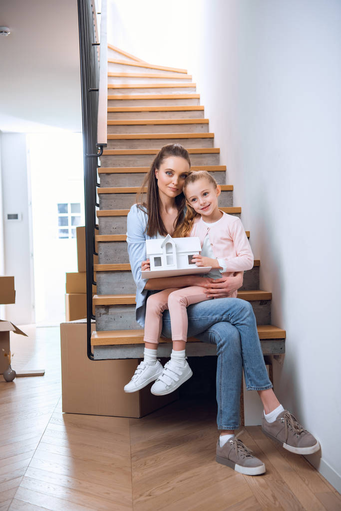 mujer feliz sentada con su hija en las escaleras y sosteniendo modelo de casa
  - Foto, Imagen