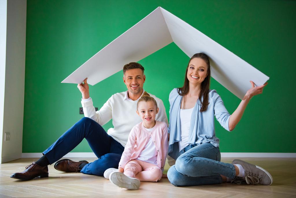 счастливые муж и жена, держа бумажную крышу, сидя на полу рядом с дочерью
 - Фото, изображение