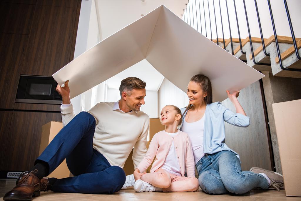 família alegre sentado sob o telhado de papel e sorrindo em nova casa
  - Foto, Imagem