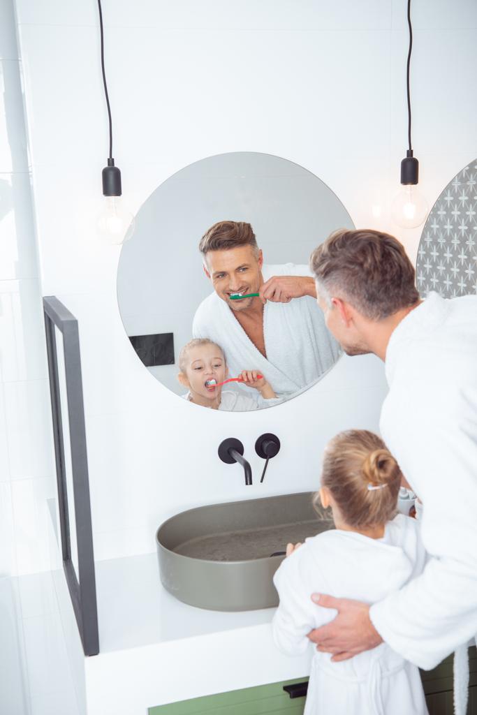 souriant père et fille brossant les dents dans les peignoirs de bain à la maison
  - Photo, image