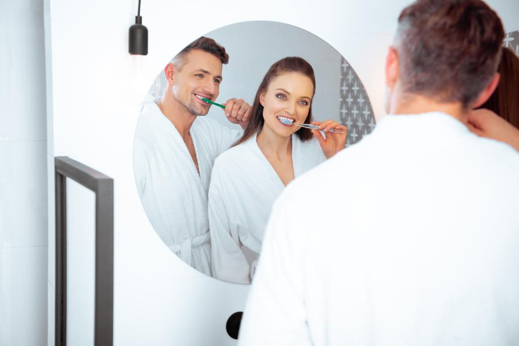 alegre pareja cepillarse los dientes y mirando en el espejo en el baño
  - Foto, Imagen
