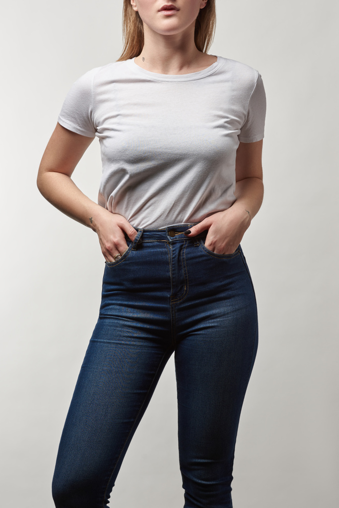 частичный вид молодой женщины в джинсах и белой футболке с копировальным пространством, изолированным на сером
 - Фото, изображение
