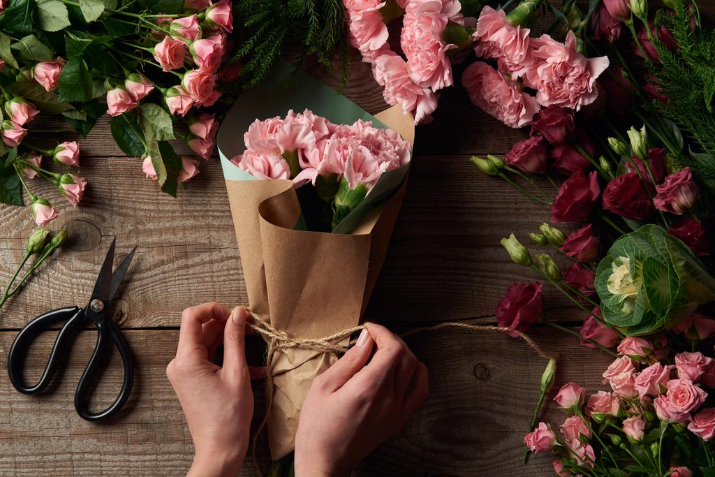 abgeschnittene Aufnahme weiblicher Hände beim Tippen von Strick auf schönem Strauß und zarten Blumen mit der Schere auf hölzerner Oberfläche - Foto, Bild