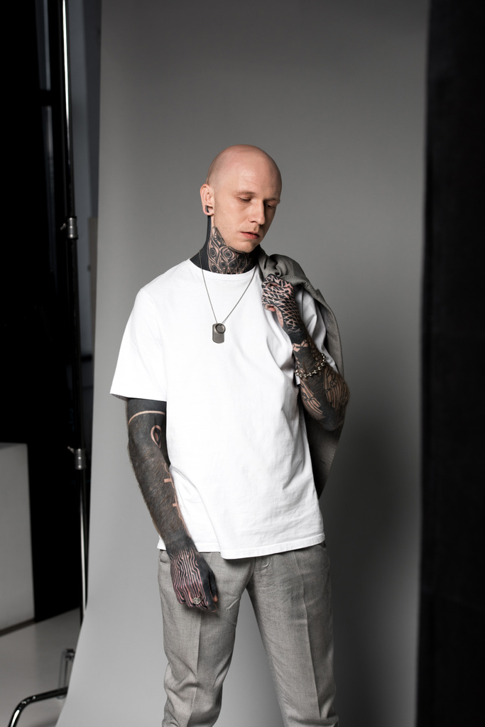 joven calvo con tatuajes sosteniendo traje chaqueta en el hombro y de pie en el estudio
 - Foto, imagen