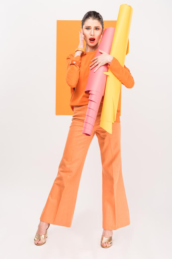 потрясенная стильная молодая женщина, держащая бумажные рулоны, трогательное лицо и позирующая с куркумой на заднем плане
 - Фото, изображение