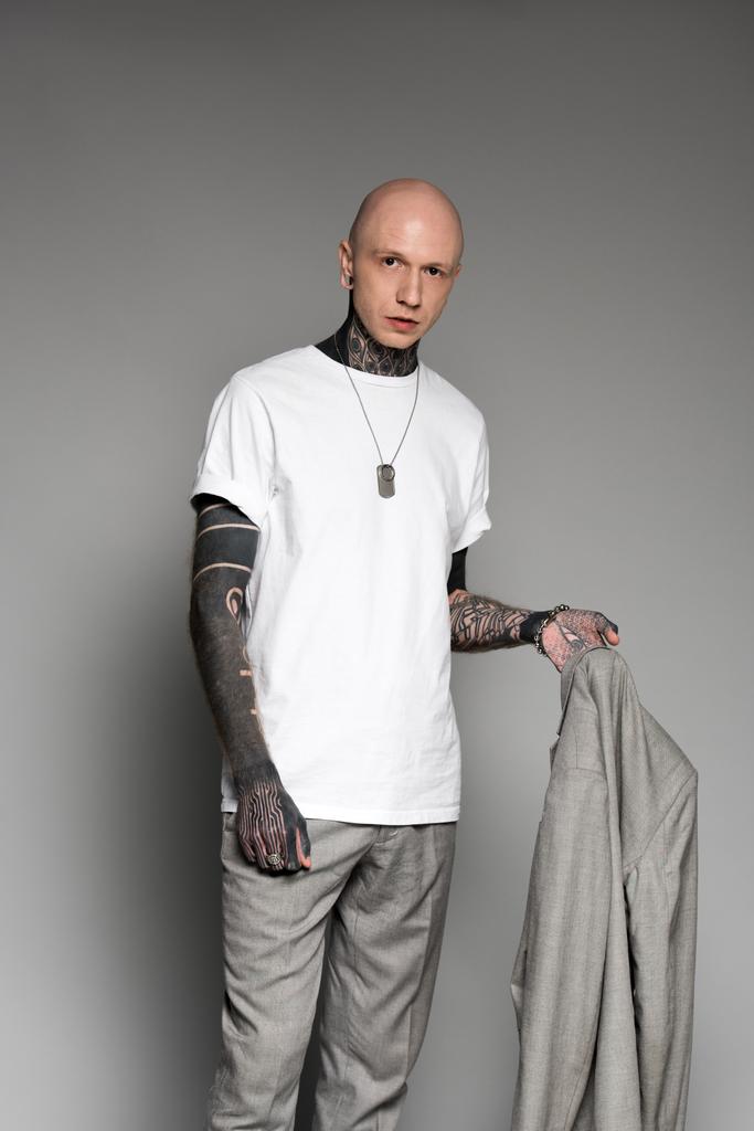 стильный лысый мужчина с татуировкой, в костюме и смотрящий в камеру на сером
  - Фото, изображение