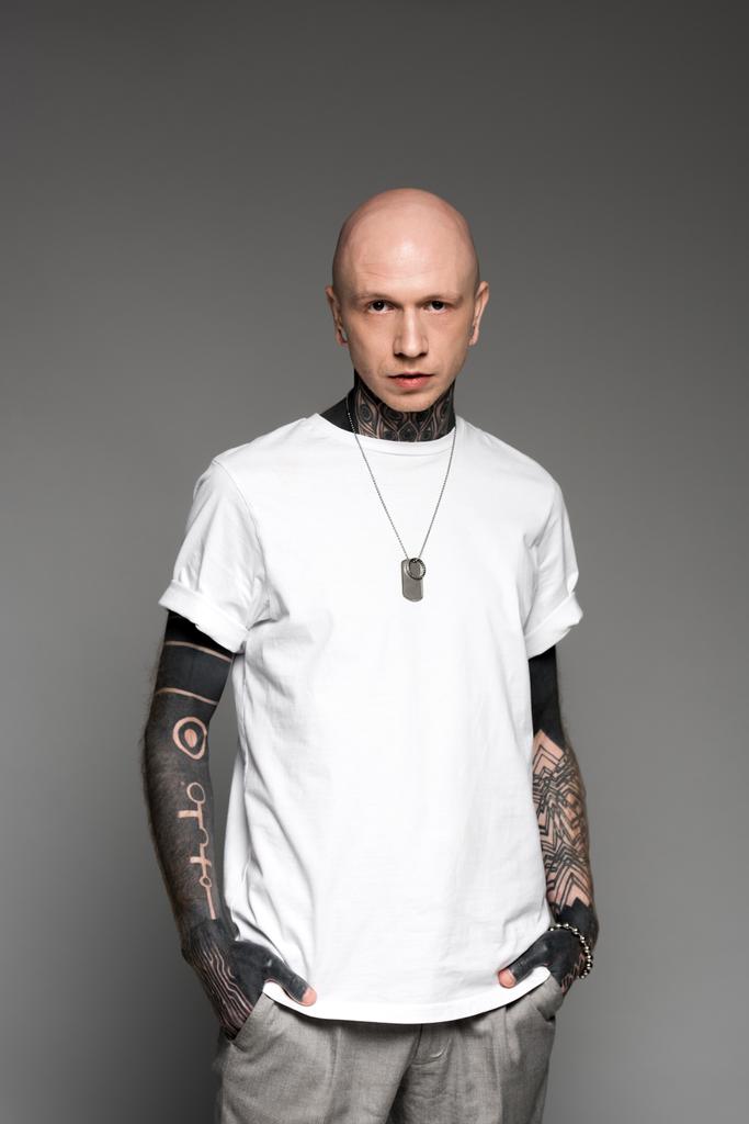 komea kalju tatuoitu mies valkoisessa t-paidassa seisoo kädet taskussa ja katselee kameraa eristettynä harmaaseen
 - Valokuva, kuva