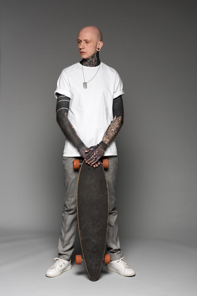 повний вигляд красивого лисого татуйованого чоловіка в білій футболці, що стоїть зі скейтбордом і дивиться на сірого
 - Фото, зображення