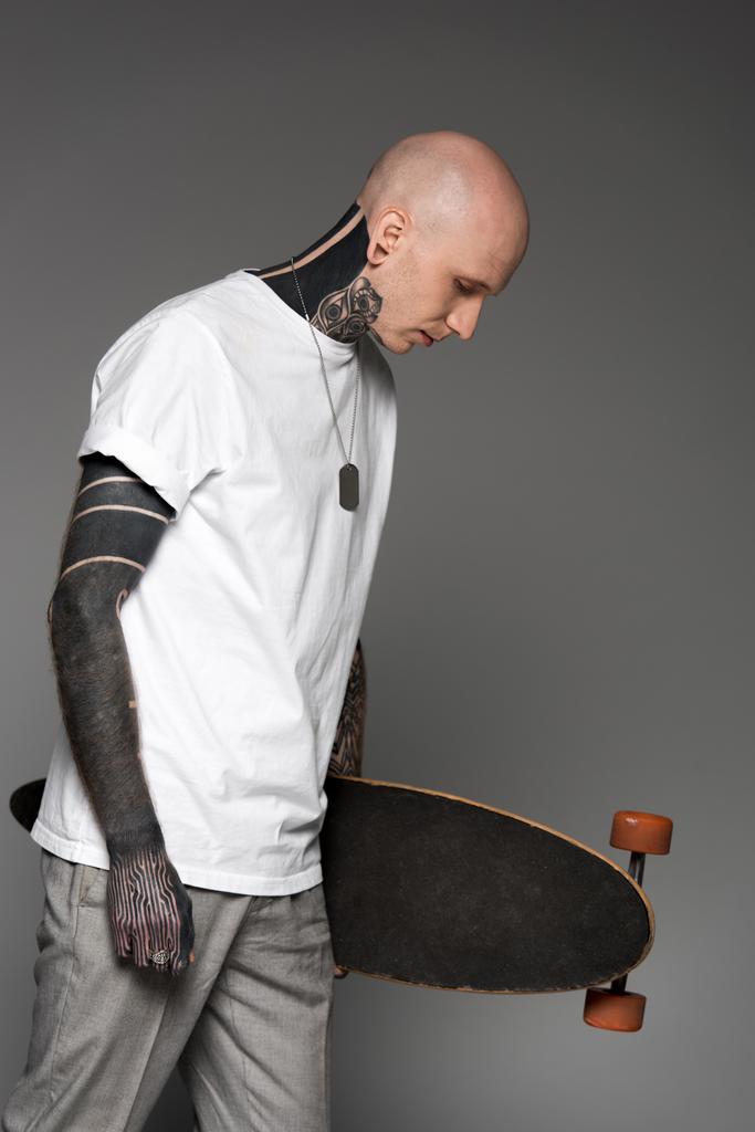 Zijaanzicht van kale getatoeëerd man in wit t-shirt holding skateboard en kijkt neer op geïsoleerde grijs - Foto, afbeelding