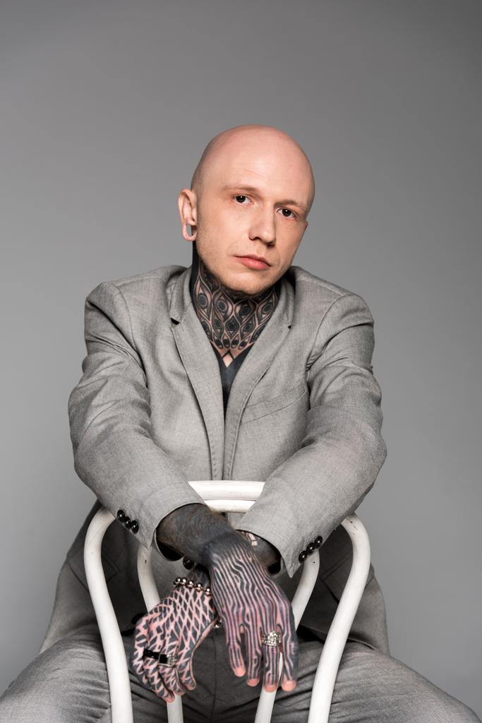 schöner stylischer glatzköpfiger tätowierter Mann im Anzug sitzt auf einem Stuhl und schaut vereinzelt in die Kamera auf grau - Foto, Bild