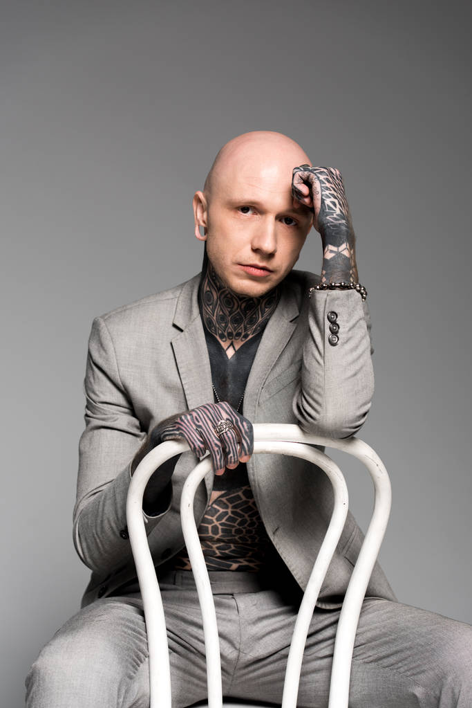 stílusos tetovált ember ül a székre, kezével a homlokát, és látszó-on fényképezőgép, elszigetelt szürke - Fotó, kép