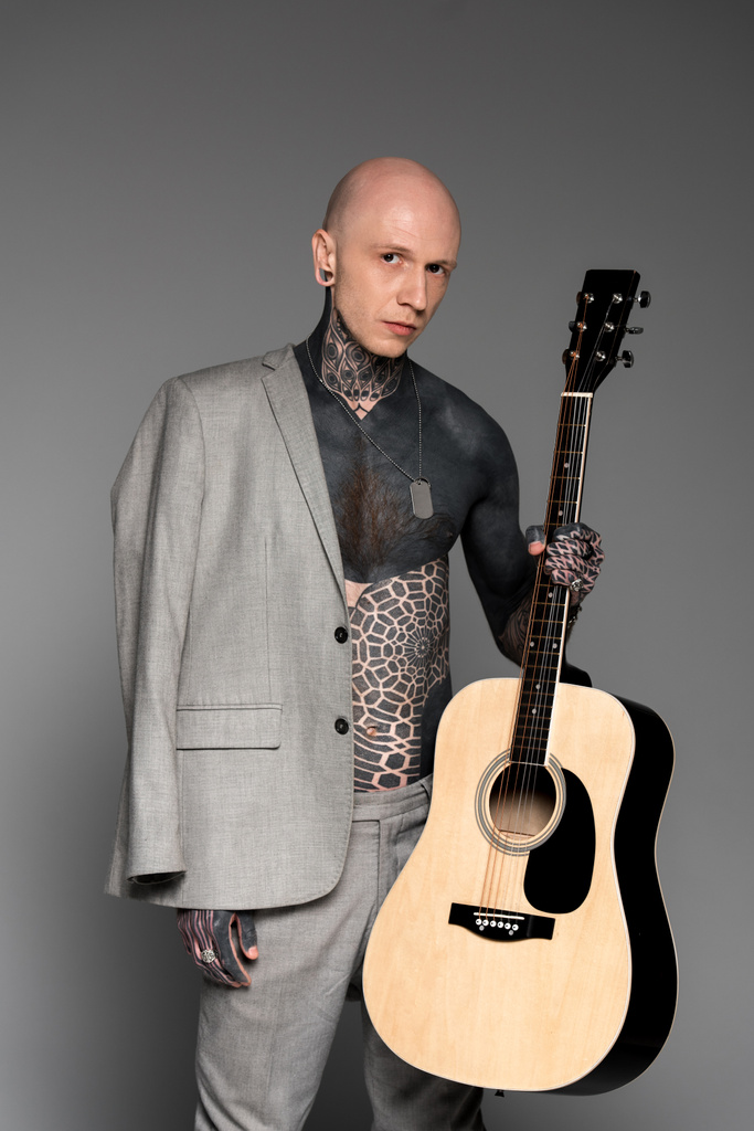 barbusig tätowierter Mann mit Anzugjacke auf der Schulter mit Akustikgitarre und Blick in die Kamera auf grau - Foto, Bild