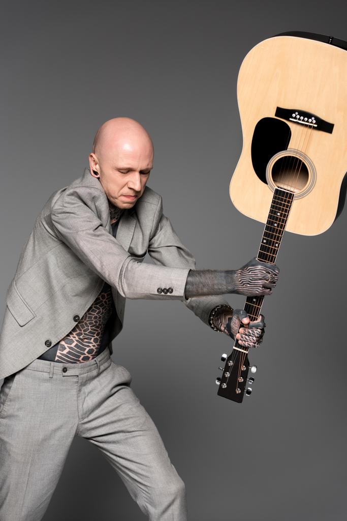 agressieve tattooed man in pak raken akoestische gitaar geïsoleerd op grijs   - Foto, afbeelding