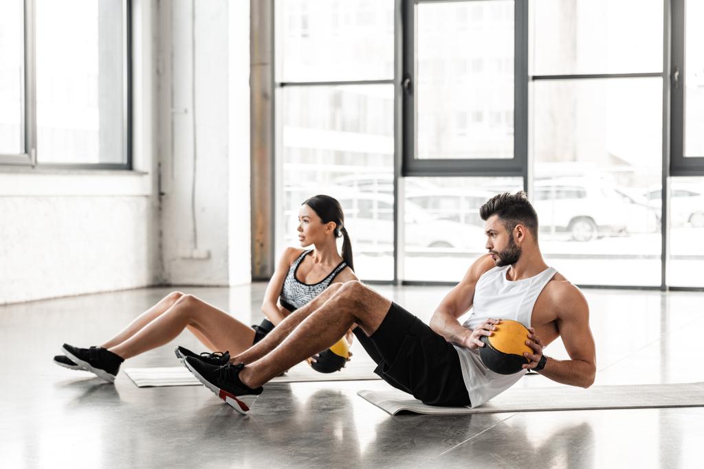 вид сбоку атлетической молодой пары в спортивной одежде, держащей медицинские мячи и занимающейся на ковриках для йоги в тренажерном зале
 - Фото, изображение